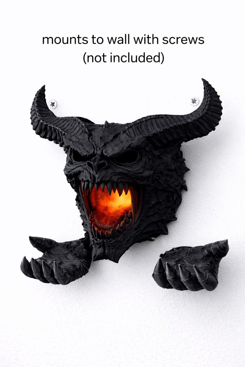 May include: A black wall-mounted sculpture of a demonic face with large horns, sharp teeth, and outstretched hands. The mouth glows with an orange light. The text "mounts to wall with screws (not included)" is above the sculpture.