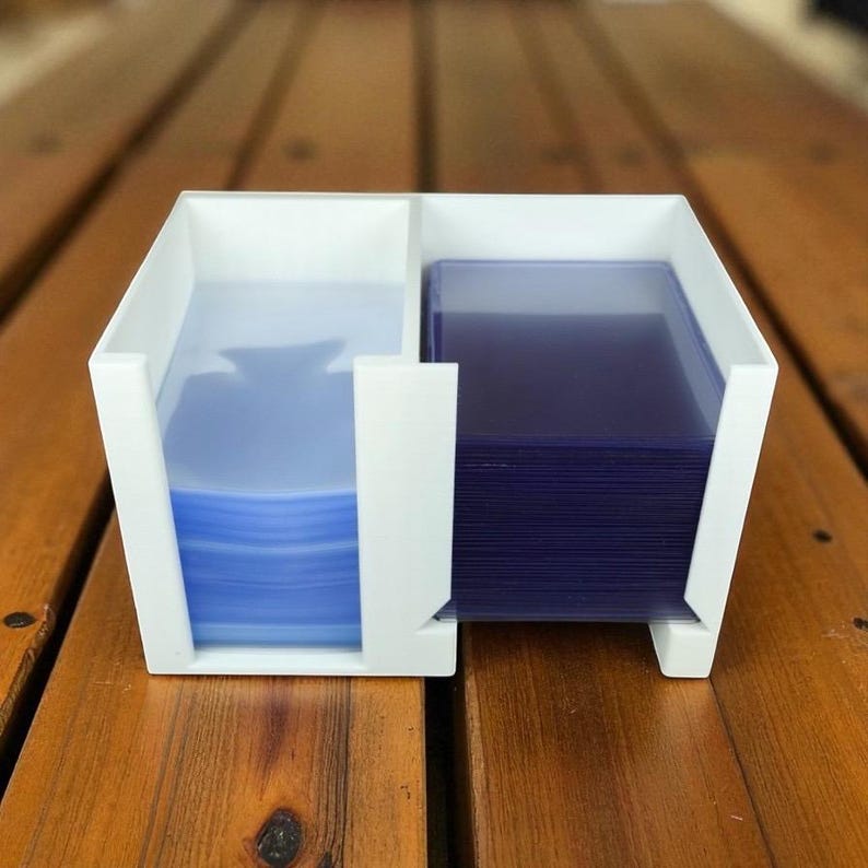 May include: A white, two-compartment card holder with stacks of blue and purple card sleeves. The holder is made of a solid, opaque material and sits on a wooden surface. The compartments are separated by a vertical divider.
