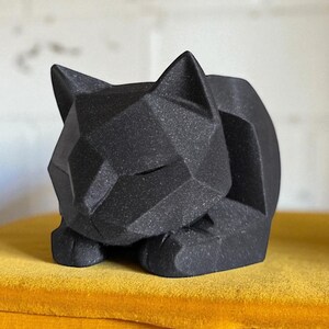 May include: A black geometric cat sculpture with a faceted design. The cat is in a resting pose, with its paws tucked beneath its body. The sculpture is displayed on a yellow surface.