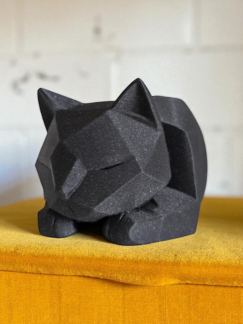 May include: A black geometric cat sculpture with a faceted design. The cat is in a resting pose, with its paws tucked beneath its body. The sculpture is displayed on a yellow surface.