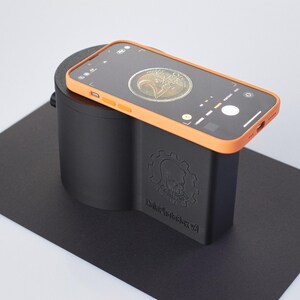 Op de afbeelding: Een zwart en oranje 3D-geprint apparaat met de tekst "Coin Scanner V2" aan de zijkant. Een smartphone met een munt in het camerabeeld is op het apparaat geplaatst.