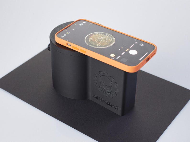 Op de afbeelding: Een zwart en oranje 3D-geprint apparaat met de tekst "Coin Scanner V2" aan de zijkant. Een smartphone met een munt in het camerabeeld is op het apparaat geplaatst.