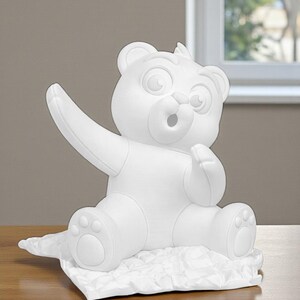 May include: A white, cartoon-style bear figurine with outstretched arm and open mouth. The bear is sitting on a textured, white surface. The figurine has large eyes and a friendly expression, suggesting a playful theme.