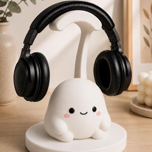 May include: A white, cartoon-style headphone stand with a smiling face and pink cheeks, holding a pair of black Sony headphones. The stand has a round base and a curved top to hold the headphones. The headphones are black.