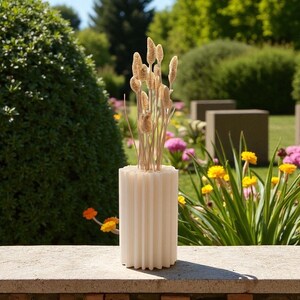 May include: A cream-colored, ribbed cylindrical vase holds dried ornamental grasses. The vase sits on a stone ledge, with a lush garden background featuring green bushes, yellow and pink flowers, and concrete structures.