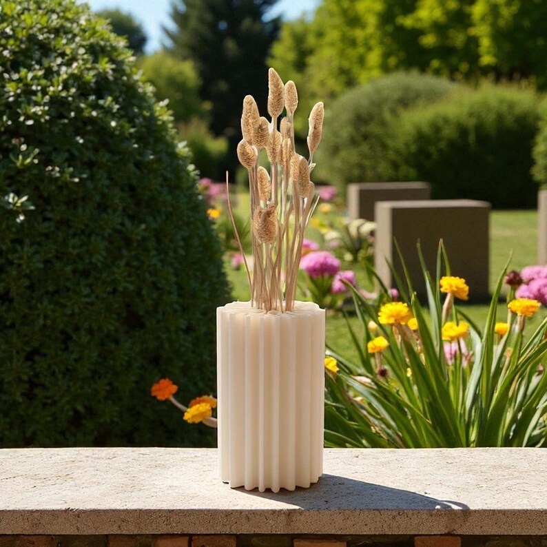 May include: A cream-colored, ribbed cylindrical vase holds dried ornamental grasses. The vase sits on a stone ledge, with a lush garden background featuring green bushes, yellow and pink flowers, and concrete structures.
