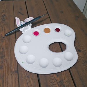 May include: A white artist's palette with a thumb hole, holding a black paintbrush and a bunny-shaped paint holder. The palette contains dabs of pink, brown, red, and green paint. The palette is on a wooden table.