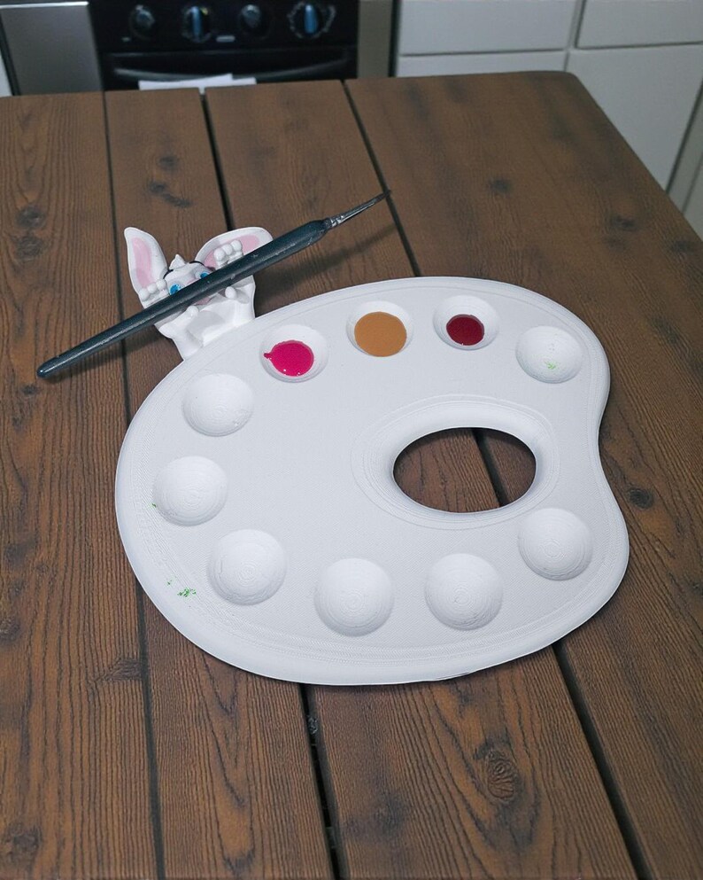 May include: A white artist's palette with a thumb hole, holding a black paintbrush and a bunny-shaped paint holder. The palette contains dabs of pink, brown, red, and green paint. The palette is on a wooden table.