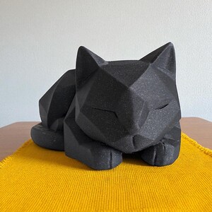 May include: A black geometric cat sculpture resting on a yellow woven placemat. The cat is in a relaxed pose with its eyes closed. The sculpture has a textured surface and is placed on a wooden table.