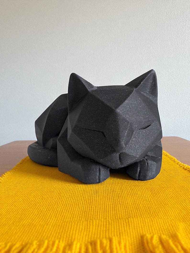 May include: A black geometric cat sculpture resting on a yellow woven placemat. The cat is in a relaxed pose with its eyes closed. The sculpture has a textured surface and is placed on a wooden table.