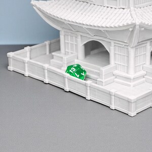 May include: A white miniature pagoda with multiple tiers and a detailed roof design. A green, multi-sided die with the number 13 is resting on a white fence in front of the pagoda. The scene is set against a gray surface and a blue background.