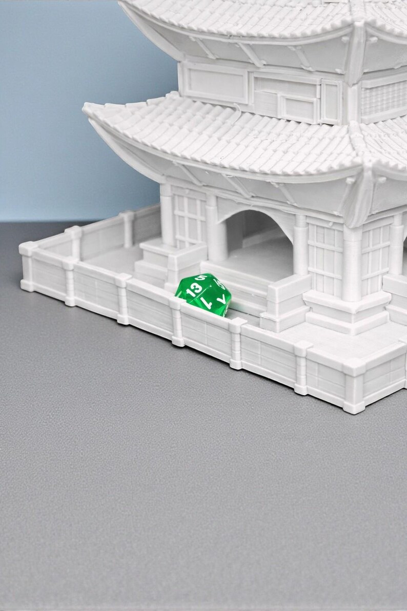 May include: A white miniature pagoda with multiple tiers and a detailed roof design. A green, multi-sided die with the number 13 is resting on a white fence in front of the pagoda. The scene is set against a gray surface and a blue background.