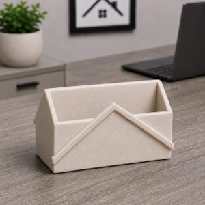 Pu&ograve; includere: Un organizer da scrivania beige a forma di casa. L'organizer ha una base rettangolare e un design a tetto spiovente. L'organizer a forma di casa &egrave; realizzato in un materiale strutturato e poggia su una scrivania di legno.