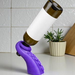 K&ouml;nnte beinhalten: Ein lila Tintenfisch-Weinflaschenhalter, der eine braune Glasflasche mit einem leeren wei&szlig;en Etikett tr&auml;gt. Der Halter steht auf einer wei&szlig;en Oberfl&auml;che, mit einer kleinen Topfpflanze und einem Holzschneidebrett im Hintergrund.