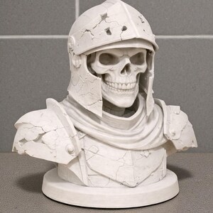 May include: A white bust of a skeleton wearing a cracked, weathered helmet and armor. The sculpture features a detailed skull and is mounted on a circular base, showcasing a gothic or fantasy theme.