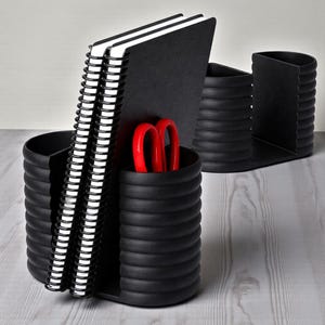 May include: A black desk organizer with a ribbed design, holding two spiral-bound notebooks, and a pair of red scissors. The organizer has multiple compartments for pens, pencils, and other office supplies. The background is a light wood grain.