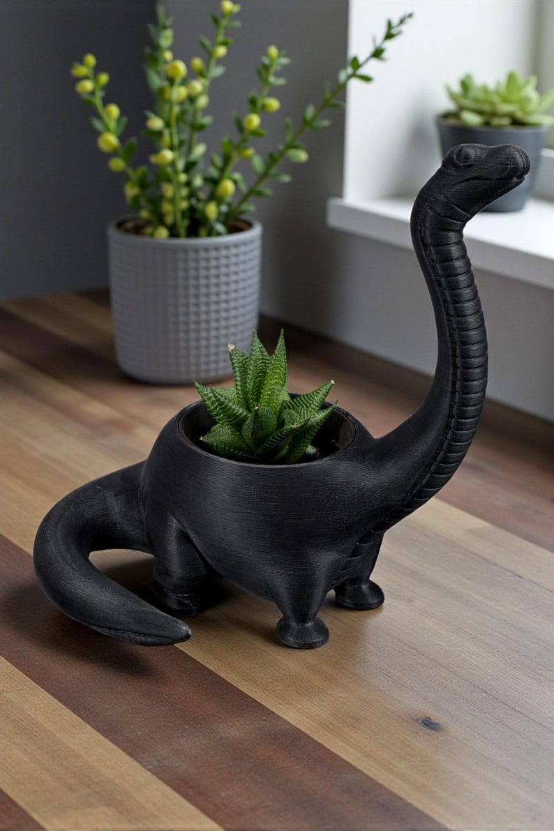 May include: Black dinosaur-shaped plant pot holding a green succulent. The pot has a long neck and a textured surface. Other potted plants are visible in the background, set on a wooden surface.