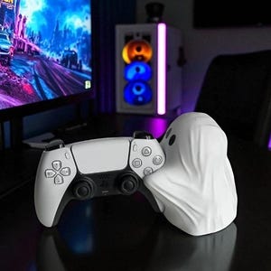 Pode incluir: Um controle de jogos branco e preto repousa sobre uma superfície preta, segurado por um suporte branco em forma de fantasma. O controle tem um design elegante com vários botões e pads direcionais. Um monitor de computador e uma torre estão ao fundo.