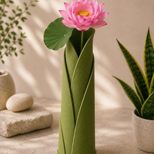 May include: A green felt vase holds a pink lotus flower with a green leaf. The vase has a unique design with overlapping panels. The background includes a plant in a pot, a stone, and a small vase.