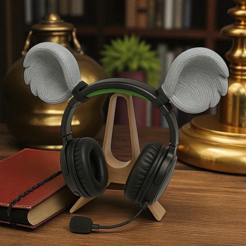 May include: Black over-ear headphones with a green accent on the headband and a flexible microphone. The headphones have gray, wing-shaped ear covers. The headphones are displayed on a wooden stand, with a book and decorative items in the background.