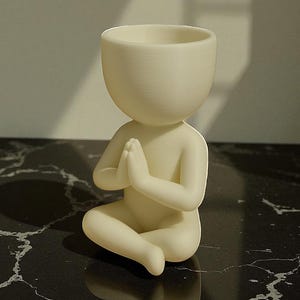 Puede incluir: Figura de color crema de una persona en posici&oacute;n de meditaci&oacute;n sentada con las manos juntas en oraci&oacute;n. La figura tiene una cabeza grande y abierta, dise&ntilde;ada para contener peque&ntilde;as plantas u objetos. La figura est&aacute; sobre una superficie negra similar al m&aacute;rmol.