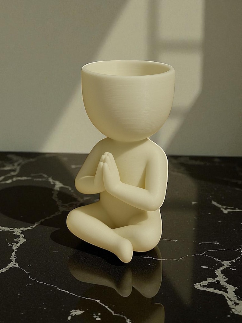 Puede incluir: Figura de color crema de una persona en posici&oacute;n de meditaci&oacute;n sentada con las manos juntas en oraci&oacute;n. La figura tiene una cabeza grande y abierta, dise&ntilde;ada para contener peque&ntilde;as plantas u objetos. La figura est&aacute; sobre una superficie negra similar al m&aacute;rmol.