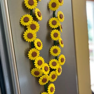 May include: A decorative sunflower garland with bright yellow petals and brown centers. The garland is arranged in a curved shape and attached to a metallic surface, likely a refrigerator. The stems are green.