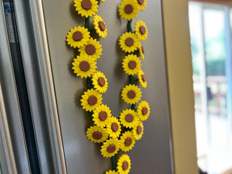 May include: A decorative sunflower garland with bright yellow petals and brown centers. The garland is arranged in a curved shape and attached to a metallic surface, likely a refrigerator. The stems are green.