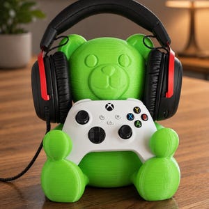 May include: A green bear-shaped headphone stand holding a white Xbox controller. Black and red gaming headphones rest on the bear's head. The stand is on a wooden surface.