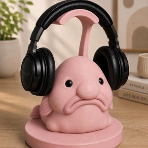 May include: A pink blob fish headphone stand with black headphones. The stand has a round base and a curved support for the headphones. The headphones are black and have the Sony logo. The stand is on a wooden surface.