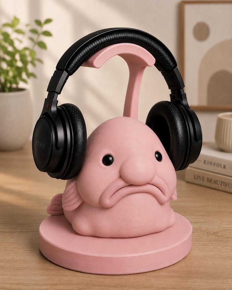 May include: A pink blob fish headphone stand with black headphones. The stand has a round base and a curved support for the headphones. The headphones are black and have the Sony logo. The stand is on a wooden surface.
