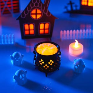 May include: A black cauldron with a glowing orange liquid inside, surrounded by three white ghost figurines and a small white candle on a blue surface.