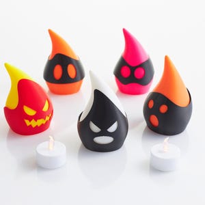 以下が含まれることがあります： カラフルで様式化されたハロウィーンをテーマにしたキャンドルホルダーのコレクション。 ホルダーは、赤、オレンジ、黒、白など、さまざまな顔のデザインと色で涙の形をしています。 2つの小さな白いティーライトキャンドルが前景にあります。