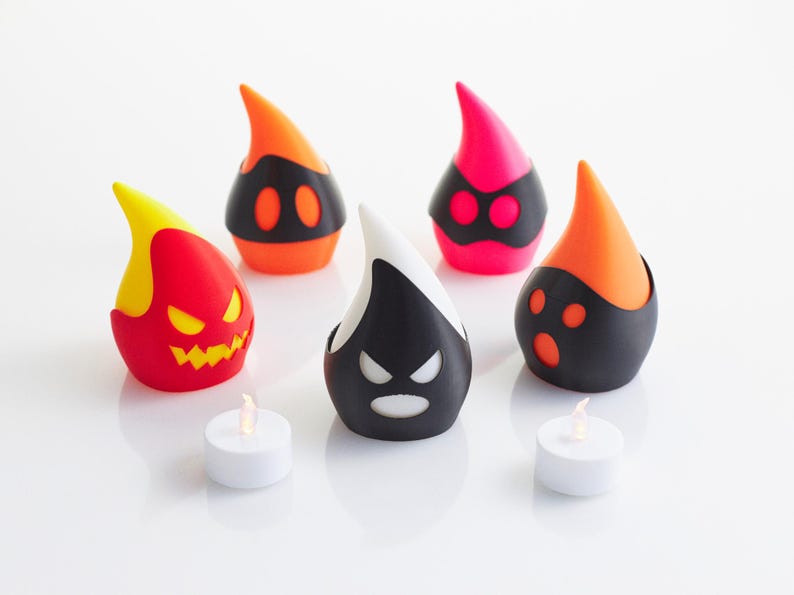 以下が含まれることがあります： カラフルで様式化されたハロウィーンをテーマにしたキャンドルホルダーのコレクション。 ホルダーは、赤、オレンジ、黒、白など、さまざまな顔のデザインと色で涙の形をしています。 2つの小さな白いティーライトキャンドルが前景にあります。