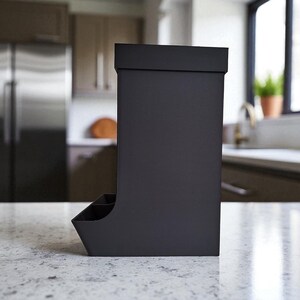 May include: A matte black kitchen utensil holder with a rectangular shape and a small compartment at the bottom. The holder is made of a solid material and sits on a speckled countertop. The background includes a stainless steel refrigerator and a window.
