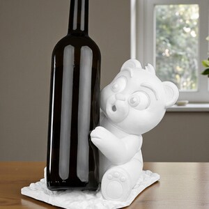 May include: A dark brown wine bottle stands next to a white bear figurine. The bear is sculpted with a surprised expression and is sitting on a textured white base. The bottle is tall and slender.