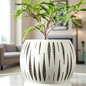 Może przedstawiać: Biała, dekoracyjna ceramiczna donica z unikalnym wzorem wycięć, zawierająca małą zieloną roślinę. Donica ma zaokrąglony kształt z pionowymi, podłużnymi otworami. Roślina ma żywe zielone liście i cienkie brązowe łodygi. Donica stoi na okrągłej podstawie.