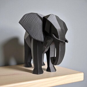 Op de afbeelding: Een geometrisch, zwart olifantbeeld met een facetontwerp. De olifant heeft grote oren, een slurf en vier poten. Het beeld staat op een lichtgekleurde houten plank tegen een grijze muur. De olifant is ongeveer 25 cm hoog.