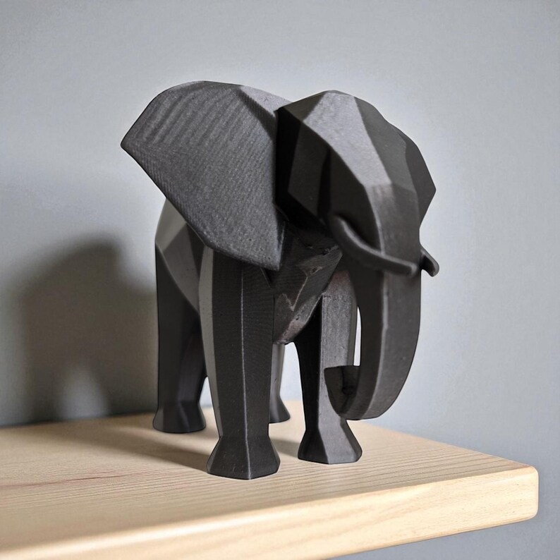 Op de afbeelding: Een geometrisch, zwart olifantbeeld met een facetontwerp. De olifant heeft grote oren, een slurf en vier poten. Het beeld staat op een lichtgekleurde houten plank tegen een grijze muur. De olifant is ongeveer 25 cm hoog.