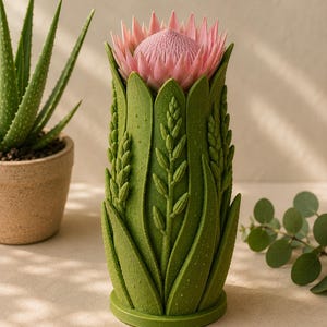 May include: A green vase with a detailed botanical design, featuring a pink protea flower. The vase has a textured surface with leaf and stem patterns. An aloe plant in a brown pot is visible in the background.