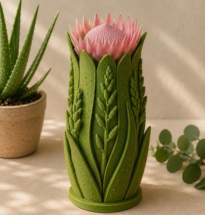 May include: A green vase with a detailed botanical design, featuring a pink protea flower. The vase has a textured surface with leaf and stem patterns. An aloe plant in a brown pot is visible in the background.