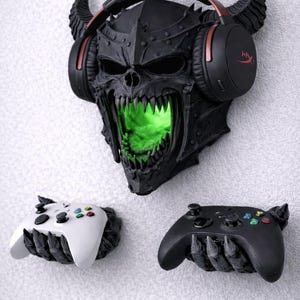 Pode incluir: Suporte de parede em forma de caveira preta com olhos verdes brilhantes e auscultadores. Dois comandos de jogo são segurados por mãos em forma de garra abaixo da caveira. Um comando é branco, o outro é preto.