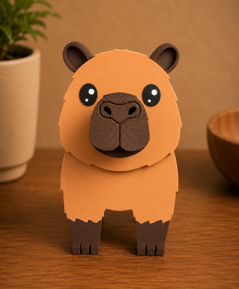 May include: A handcrafted, three-dimensional capybara figure. The figure is primarily orange with dark brown accents on the ears, nose, and feet. The capybara has large, round eyes with white highlights. The figure is standing on a wooden surface.