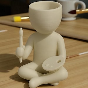 May include: A cream-colored figurine of a person sitting cross-legged, holding a paintbrush and a palette. The figure has a large, open head, possibly for use as a planter or container. The figurine is on a wooden surface.
