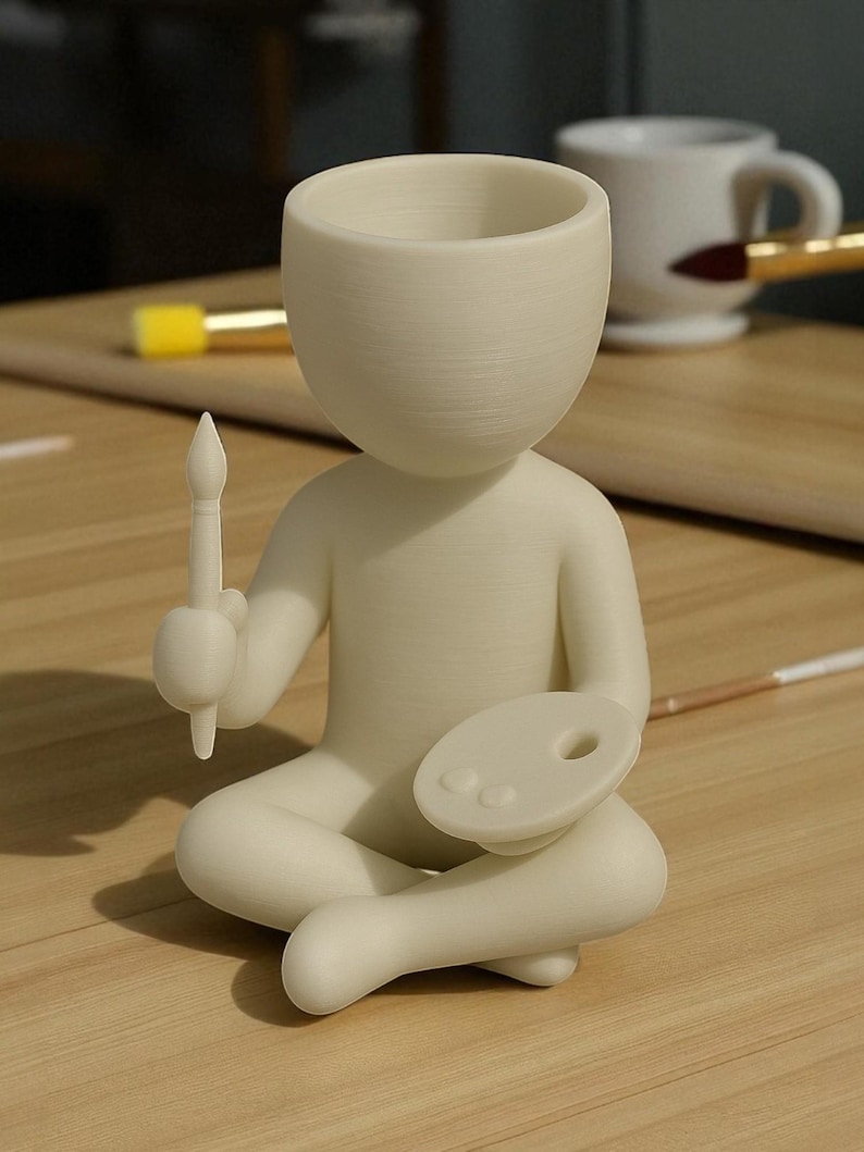 May include: A cream-colored figurine of a person sitting cross-legged, holding a paintbrush and a palette. The figure has a large, open head, possibly for use as a planter or container. The figurine is on a wooden surface.