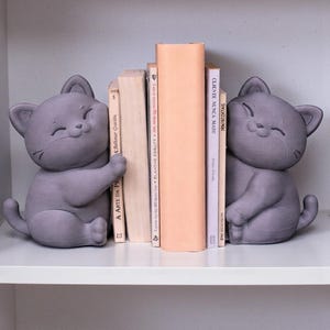 May include: Two gray cat-shaped bookends sit on a white shelf, flanking a stack of books. The bookends have a smiling face and are holding the books. The books have titles in Portuguese and French.