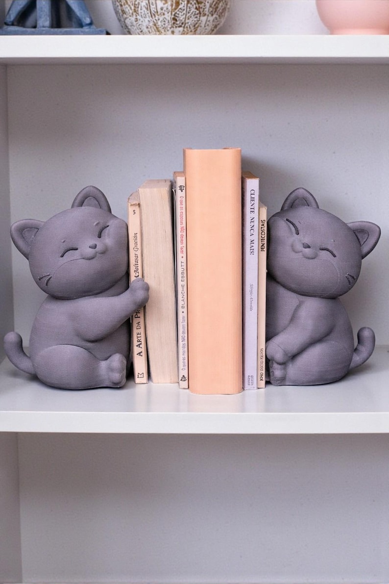 May include: Two gray cat-shaped bookends sit on a white shelf, flanking a stack of books. The bookends have a smiling face and are holding the books. The books have titles in Portuguese and French.