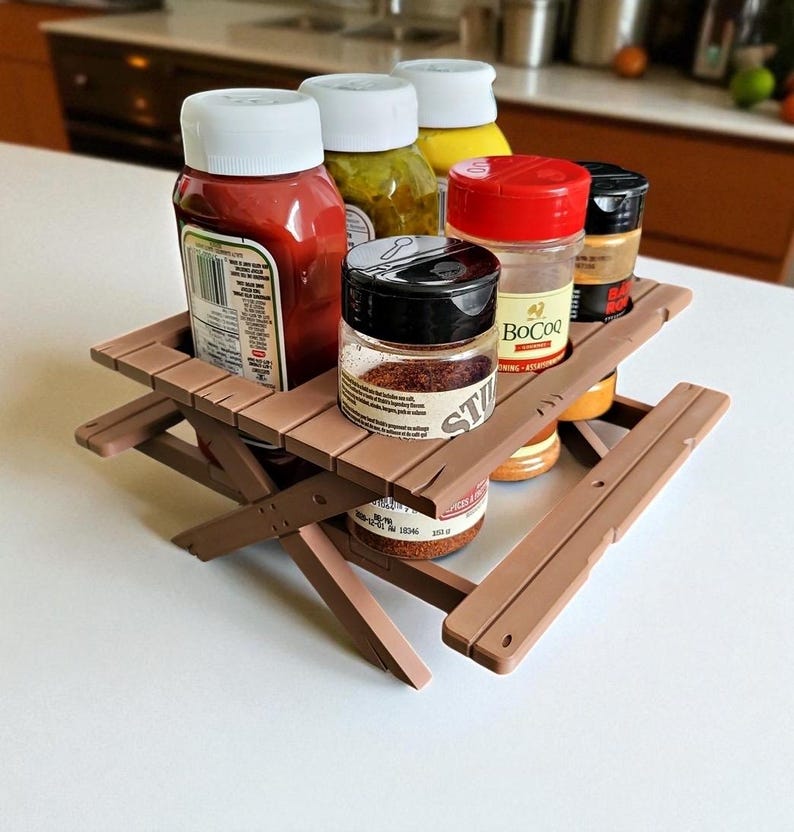 Puede incluir: Una peque&ntilde;a mesa de picnic marr&oacute;n sostiene varias botellas de condimentos y frascos de especias. Las botellas incluyen ketchup, mostaza y otras salsas. Los frascos contienen especias. La mesa es de color marr&oacute;n.