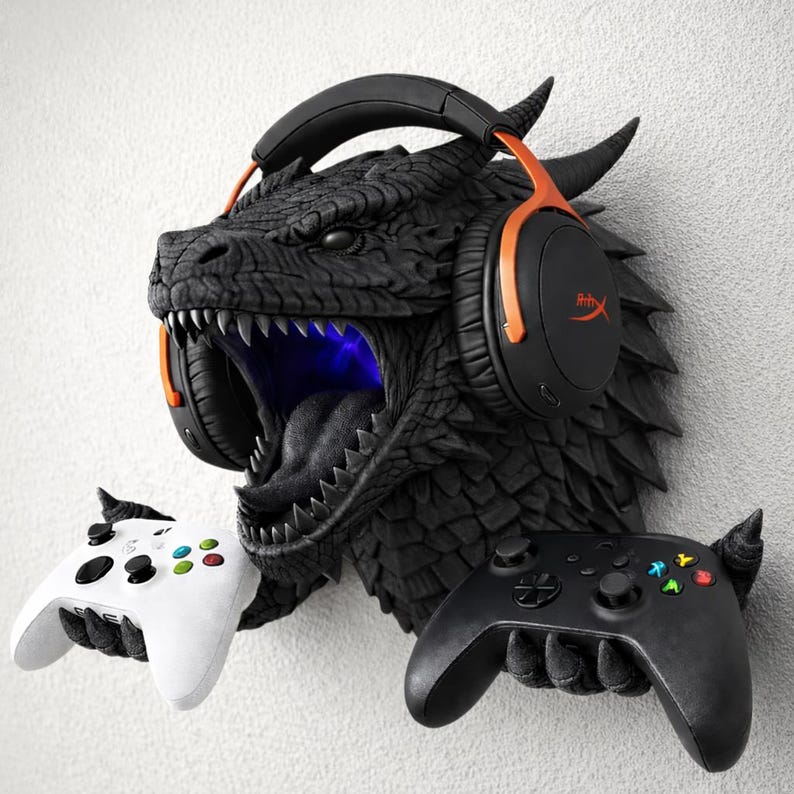 May include: A black dragon head wall mount with orange and black headphones and two game controllers. The dragon's mouth is open, revealing sharp teeth and a blue light. One white and one black game controller are held in the dragon's hands.