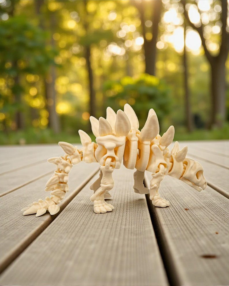 May include: A cream-colored, articulated dinosaur skeleton model. The model features a detailed design with visible ribs, a spiky back, and a long tail. It is posed on a wooden surface with a blurred background of trees.